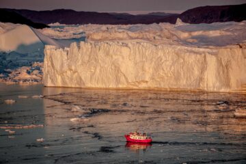 Rotes Schiff hebt sich vor großer Eiswand ab in orangenem Licht