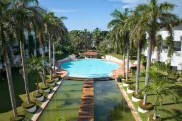 Blick auf eine Pool-Anlage eines Hotels, mit Palmen und Steg zum Pool.