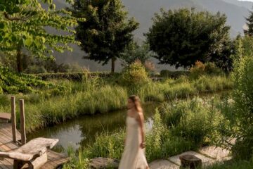 Frau in weißem Kleid, geht an einem Teich vorbei, im Hintergrund Berge.